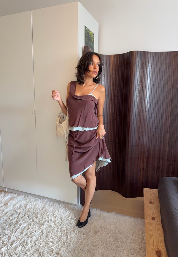 Mujer con vestido marrón con ribete claro, de pie sobre una alfombra blanca cerca de un armario y una pantalla plegable de madera en una habitación suavemente iluminada.