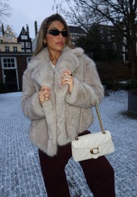 Γυναίκα που φοράει γκρι παλτό από γούνα, σκούρα γυαλιά ηλίου, πολλά δαχτυλίδια και κρατάει μια λευκή καπιτονέ τσάντα με χρυσή αλυσίδα σε εξωτερικό χώρο σε χιονισμένο λιθόστρωτο δρόμο.