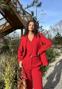 Sarkans pielāgots kostīms ar īsu žaketi un augsta vidukļa biksēm. Audums šķiet gluds ar matētu apdari. Brūna somiņa ar kniedēm.