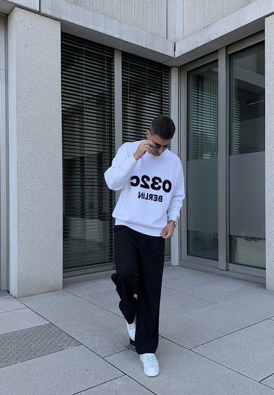 Sudadera blanca con el texto gráfico negro "0320 BERLÍN," combinada con pantalones holgados negros y zapatillas blancas, frente a una arquitectura moderna.