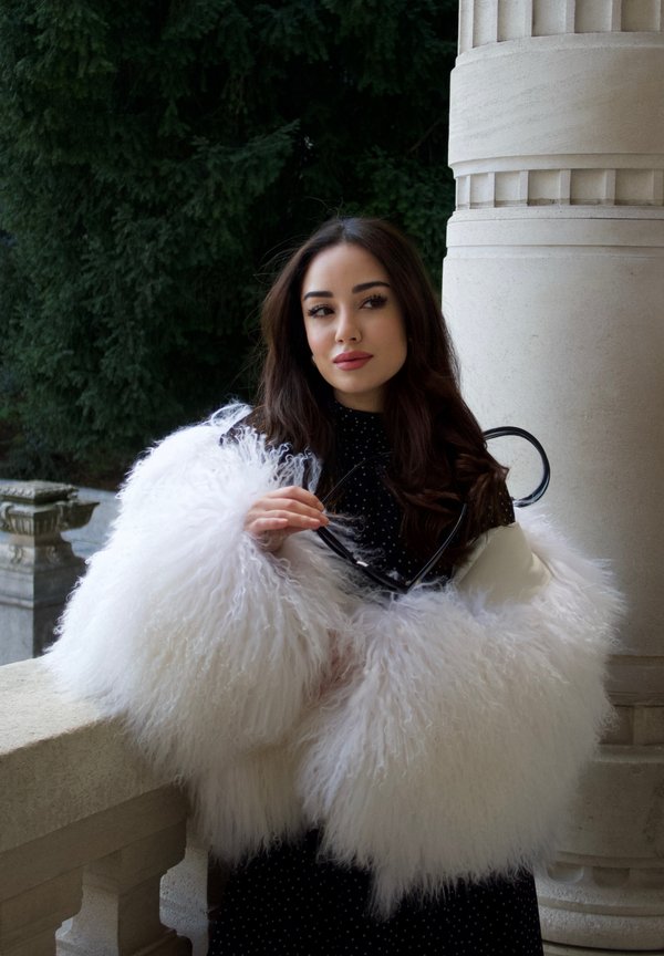 Jeune femme aux longs cheveux noirs portant un manteau blanc duveteux et une robe noire à pois, tenant des lunettes de soleil près d'une colonne en pierre.