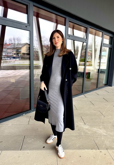 Abrigo negro de doble botonadura, vestido gris ajustado, leggings negros y zapatillas blancas con detalles en marrón. Bolso negro con herrajes dorados.