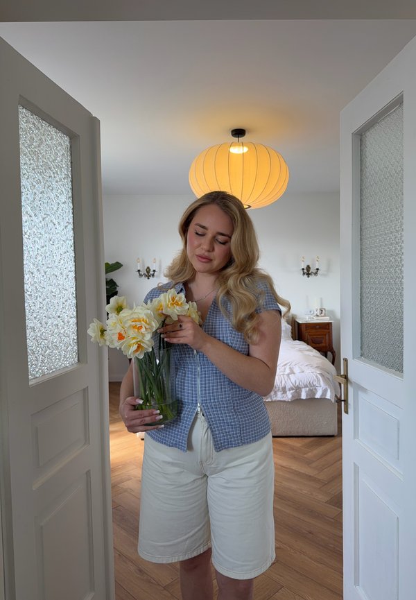 Frau in blauem Hemd und weißen Shorts hält eine Vase mit gelben und weißen Blumen und steht in einem Türrahmen eines Schlafzimmers mit Holzboden.
