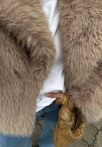 Világosbarna műszőrméből készült kabát, puha textúrával, fehér inggel és világoskék farmerrel párosítva. A kéz egy barna fonott clutch-ot tart.