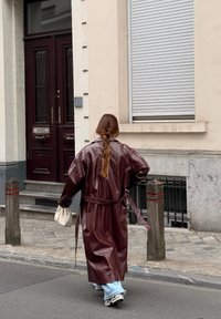 Bordeaux leren lange jas met brede mouwen en ceintuur, gecombineerd met lichtblauwe jeans en sneakers. Straatscène met straatstenen pad.