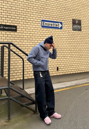 Veste à rayures bleu et blanc avec capuche, pantalon large noir, et baskets roses. Debout près d'un mur en briques avec des panneaux directionnels.
