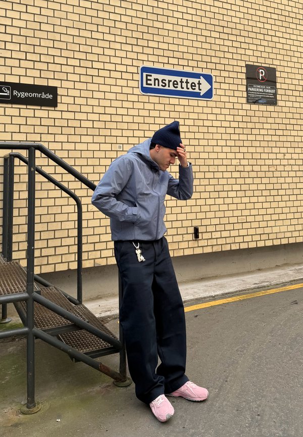 Blue and white striped jacket with hood, black wide-leg pants, and pink sneakers. Standing near a brick wall with directional signs.