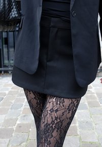 Črn krojen blazer čez oprijet črn zgornji del, kombiniran s kratko črno krilo in črnimi pajkicami z vzorcem čipke; vidni teksturni detajli.