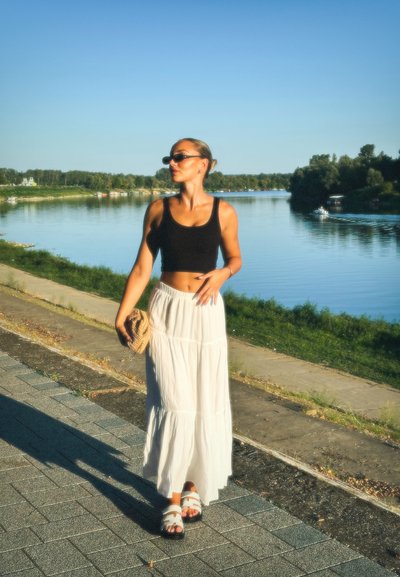 Top corto negro, falda larga blanca de volantes, sandalias blancas con tiras. Los accesorios incluyen gafas de sol y un bolso de paja beige. Fondo de un río sereno.