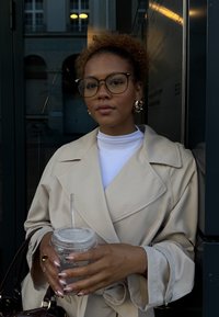 Kvinde med krøllet hår og briller iført en beige trenchcoat holder en plastkop med et sugerør og står nær en bygningsindgang.