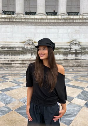 Joven sonriente, vestida con una camiseta negra de hombros descubiertos y una gorra, de pie sobre un suelo de piedra con patrón frente a un gran edificio antiguo con columnas.