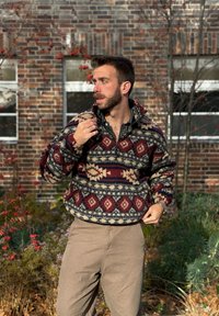 Fleece-pullover i bordeaux, creme og marineblå med geometriske mønstre, halvlange lynlås og elastiske manchetter. Bæres med beige bukser.