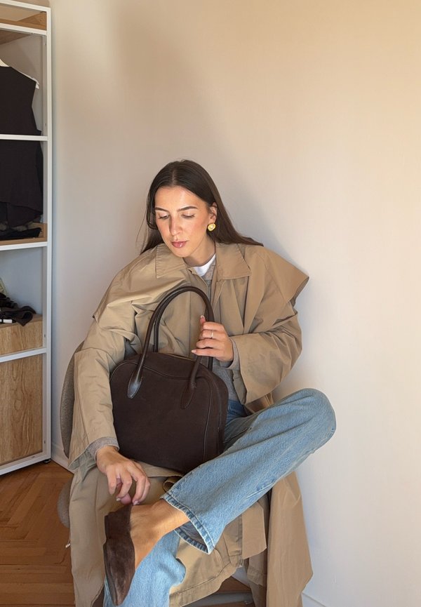 Bolsa de couro castanha com alças de ombro, usada com um casaco oversized bege, jeans azuis e sapatos castanhos de slip-on. Brincos amarelos visíveis.