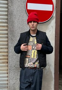 Jeune homme portant un bonnet rouge, un pull à motifs, un gilet noir et un pantalon rayé, debout sous un panneau de circulation d'interdiction d'entrée contre un mur en béton.
