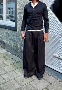 Homme portant une chemise henley noire à manches longues et un large pantalon noir, debout sur un trottoir en pierre devant un mur en briques grises et une porte blanche.