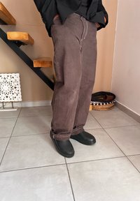 Καφέ, φαρδιά παντελόνια με ορατές ραφές και μαύρα υποδήματα με υφημένο πάτο, σε ένα πλακιδωτό εσωτερικό.