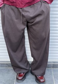 Løse, brune bukser laget av et glatt stoff, med elastisk midje og sidelommer, kombinert med skinnende, mørkebrune sko.