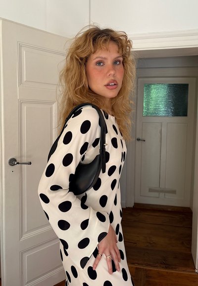Vestido negro y blanco con lunares, de manga larga y tejido texturizado, combinado con un pequeño bolso negro. Suelo de madera natural y puerta de color claro.