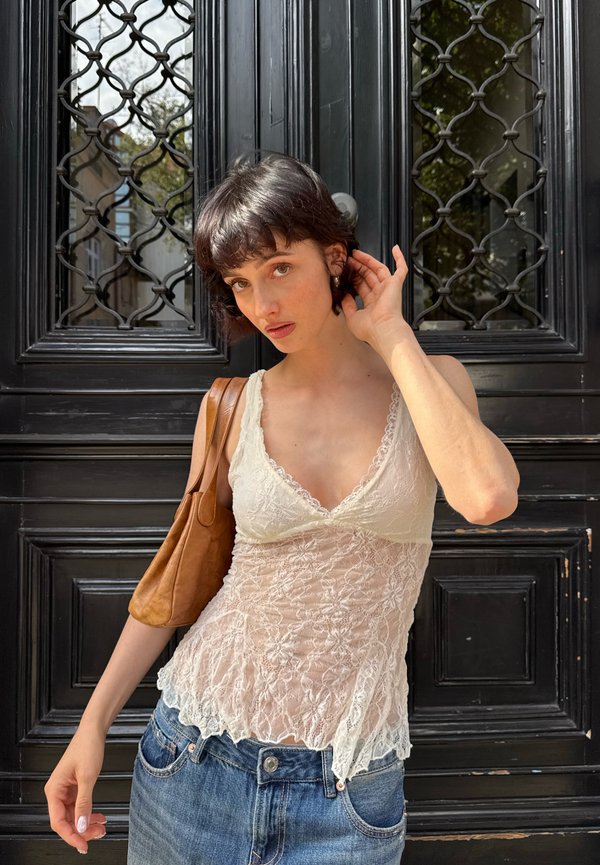 Joven mujer con el pelo corto y oscuro, vistiendo una blusa blanca de encaje y vaqueros azules, de pie frente a una puerta negra ornamentada, llevando un bolso marrón al hombro.
