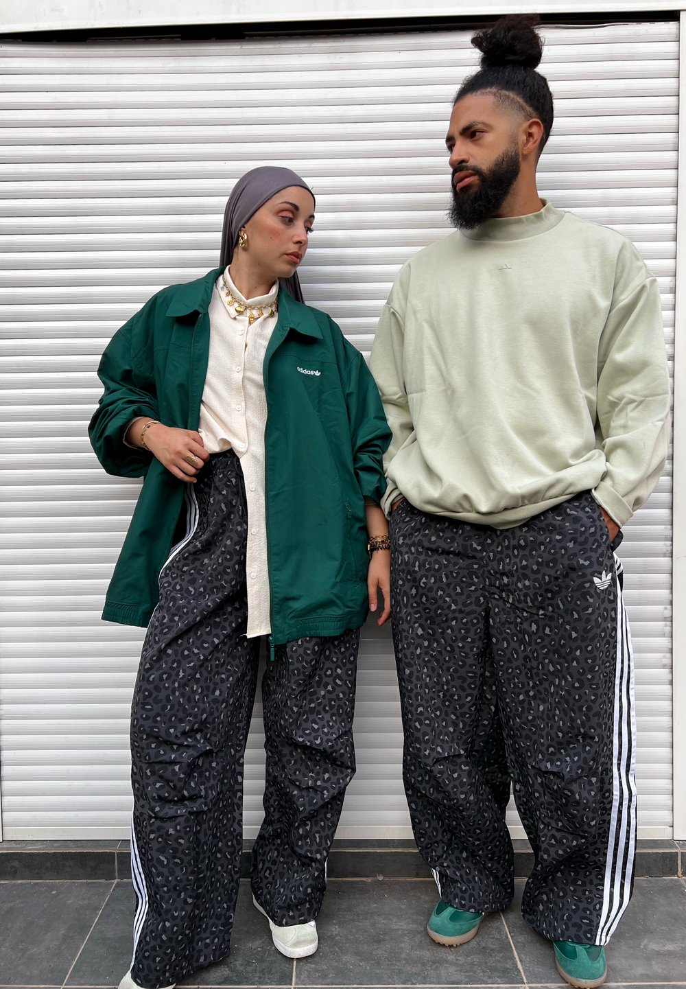 Duas pessoas vestindo roupas casuais. A mulher usa um casaco verde oversized, uma camisa creme e calças com estampa de leopardo. O homem veste um casaco leve verde claro e calças estampadas com riscas brancas.