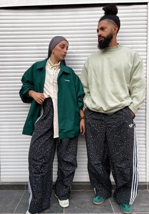Dos personas vestidas con ropa casual. La mujer lleva una chaqueta verde de gran tamaño, una camisa color crema y pantalones con estampado de leopardo. El hombre viste una sudadera verde clara y pantalones estampados con rayas blancas.