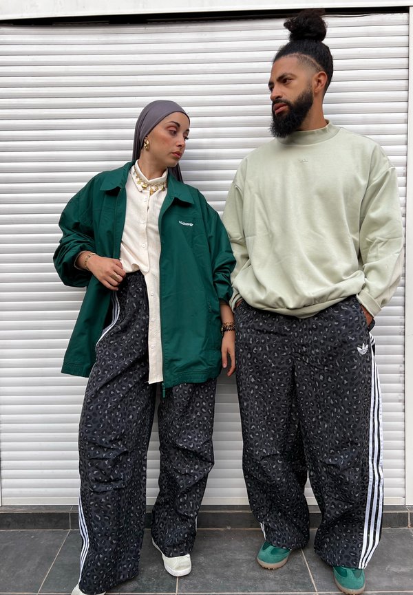 Duas pessoas vestindo roupas casuais. A mulher usa um casaco verde oversized, uma camisa creme e calças com estampa de leopardo. O homem veste um casaco leve verde claro e calças estampadas com riscas brancas.