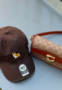 Bruine baseballcap met geborduurd corgi en designer tan handtas met bruine leren afwerking op een grijze ondergrond.