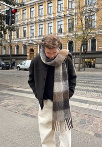 A man wearing a black coat, a large checked scarf, and white trousers stands on a city pavement beneath a red traffic light, with a historic building in the background.