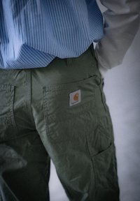 Pantaloni cargo verzi cu buzunare mari și etichetă Carhartt. Purtați cu o cămașă cu dungi albastre deschise, parțial ascunsă în imagine.