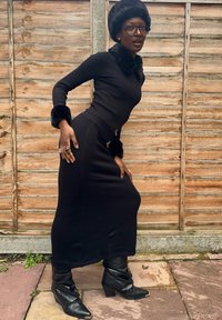 Čierny rebrovaný top s dlhým rukávom a kožušinovým golierom, čierna pletená sukňa a čierne kožené čižmy po kolená, doplnené prsteňmi a okuliarmi.