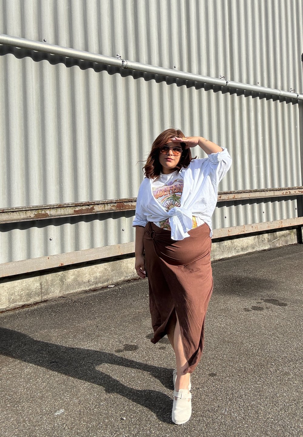 Bruine midi rok met een zijsplit, witte strikblouse over een graphic t-shirt, en lichtgekleurde instappers. Zonnebril wordt gedragen.