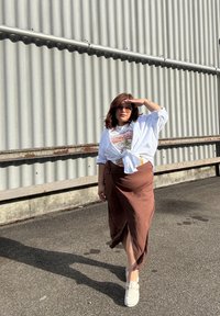 Bruine midi rok met een zijsplit, witte strikblouse over een graphic t-shirt, en lichtgekleurde instappers. Zonnebril wordt gedragen.
