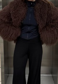 Veste en fausse fourrure marron à texture bouclée, associée à un haut noir ajusté et un pantalon noir à jambes larges. Le fond présente un cadre simple d'ascenseur.