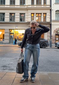 Personne portant des lunettes de soleil, un cardigan foncé et un jean ample, tenant un sac fourre-tout à motifs, debout sur un trottoir en ville près de voitures garées.