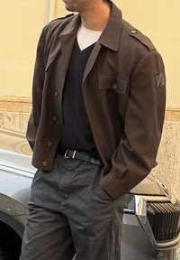 Homme portant une veste marron foncé, un pull noir sur une chemise blanche, un pantalon gris, debout les mains dans les poches près d'une voiture noire.