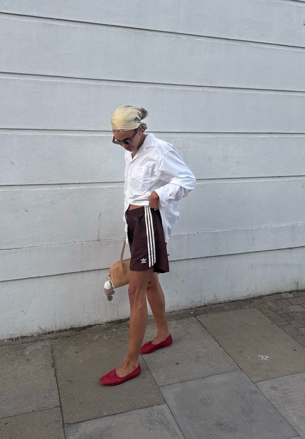 Persona con camisa blanca, pantalones cortos oscuros con rayas, zapatos rojos y pañuelo en la cabeza, de pie en una acera frente a una pared gris.