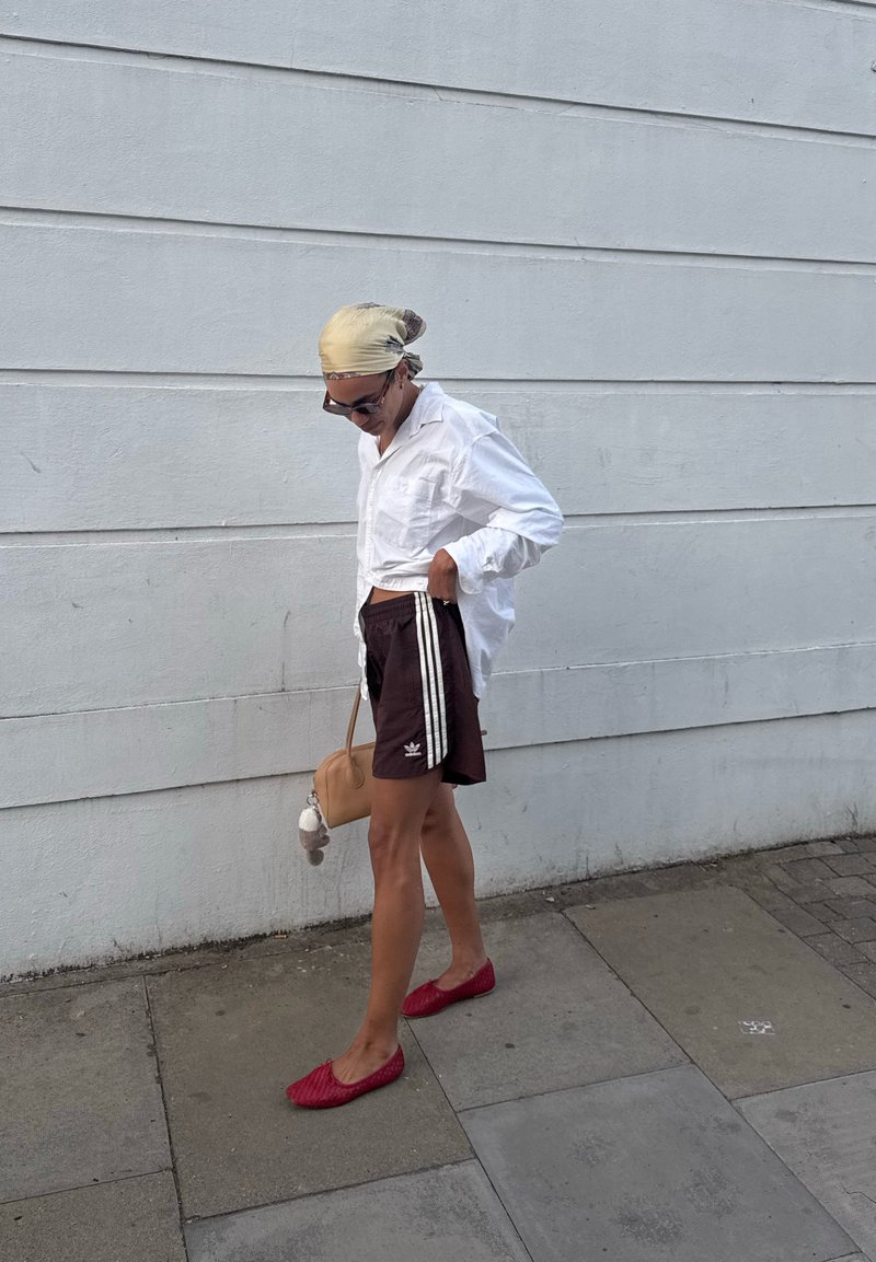 Personne portant une chemise blanche, un short foncé avec des rayures, des chaussures rouges et un foulard sur la tête, debout sur un trottoir devant un mur gris.