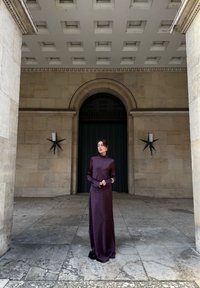 Abito lungo in raso viola scuro con maniche lunghe e collo alto; texture liscia. Ambientato davanti a un muro di pietra con motivi geometrici e lanterne.