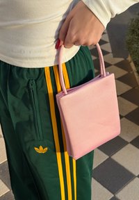 Une main tient un petit sac à main rose au-dessus d'un pantalon de survêtement Adidas vert avec des rayures jaunes, debout sur des carreaux extérieurs à damiers.