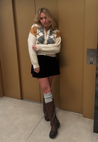 Πλεκτό κρεμ πουλόβερ με πολύχρωμα γεωμετρικά σχέδια, μαύρη φούστα, καφέ μπότες μέχρι το γόνατο και γκρι κάλτσες με ribbed υφή.