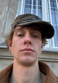 En person med lys brunt hår bærer en camouflage-mønstret kasket og en brun jakke. Sølv øreringe i form af hoops ses i begge ører.