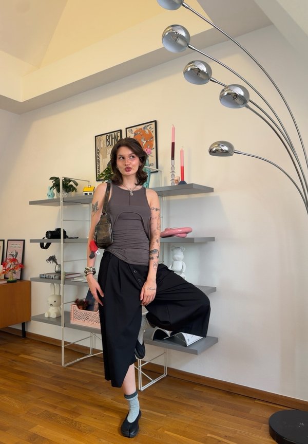 Junge Frau mit Tattoos posiert drinnen, trägt ein ärmelloses Oberteil und weite schwarze Hosen, steht auf einem Bein nahe Regalen und einer Stehlampe.