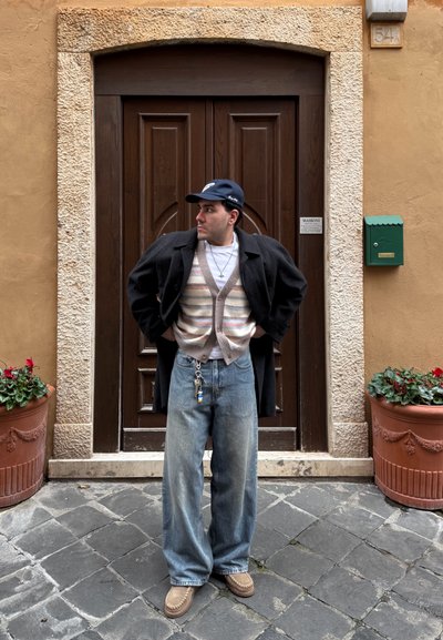 Hombre con cárdigan de rayas, abrigo negro, gorra azul y pantalones vaqueros anchos de pie sobre adoquines frente a una puerta de madera oscura enmarcada por piedra texturizada.