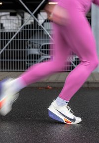 Corridore indossando leggings rosa e scarpe da ginnastica Nike bianche a metà falcata su una strada asfaltata vicino a una recinzione metallica.