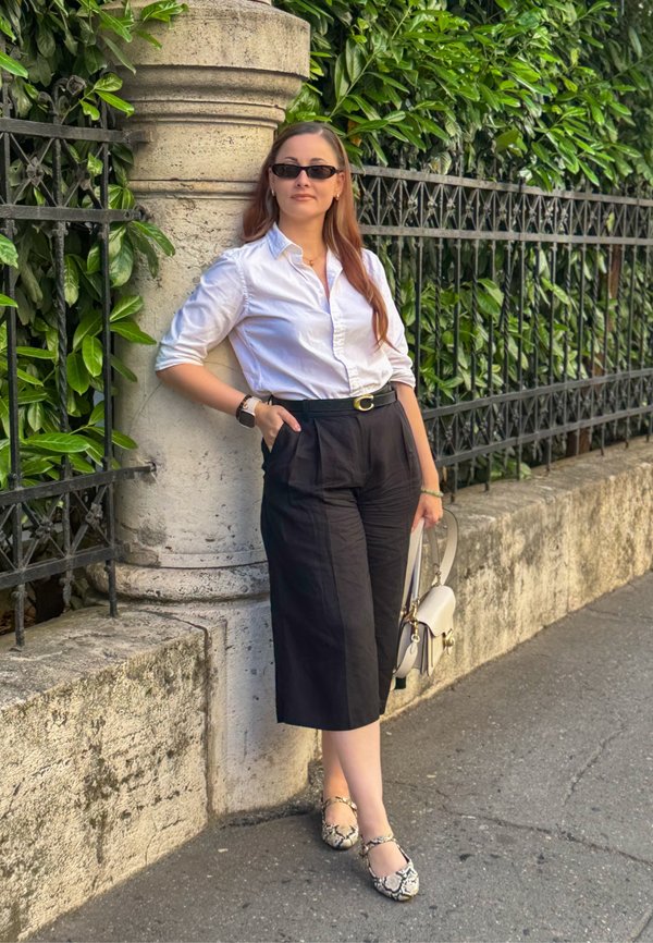Frau mit Sonnenbrille, weißem Hemd, schwarzen weiten Hosen und gemusterten Schuhen lehnt an einer steinernen Säule mit grüner Bepflanzung und einem Eisenzaun dahinter.