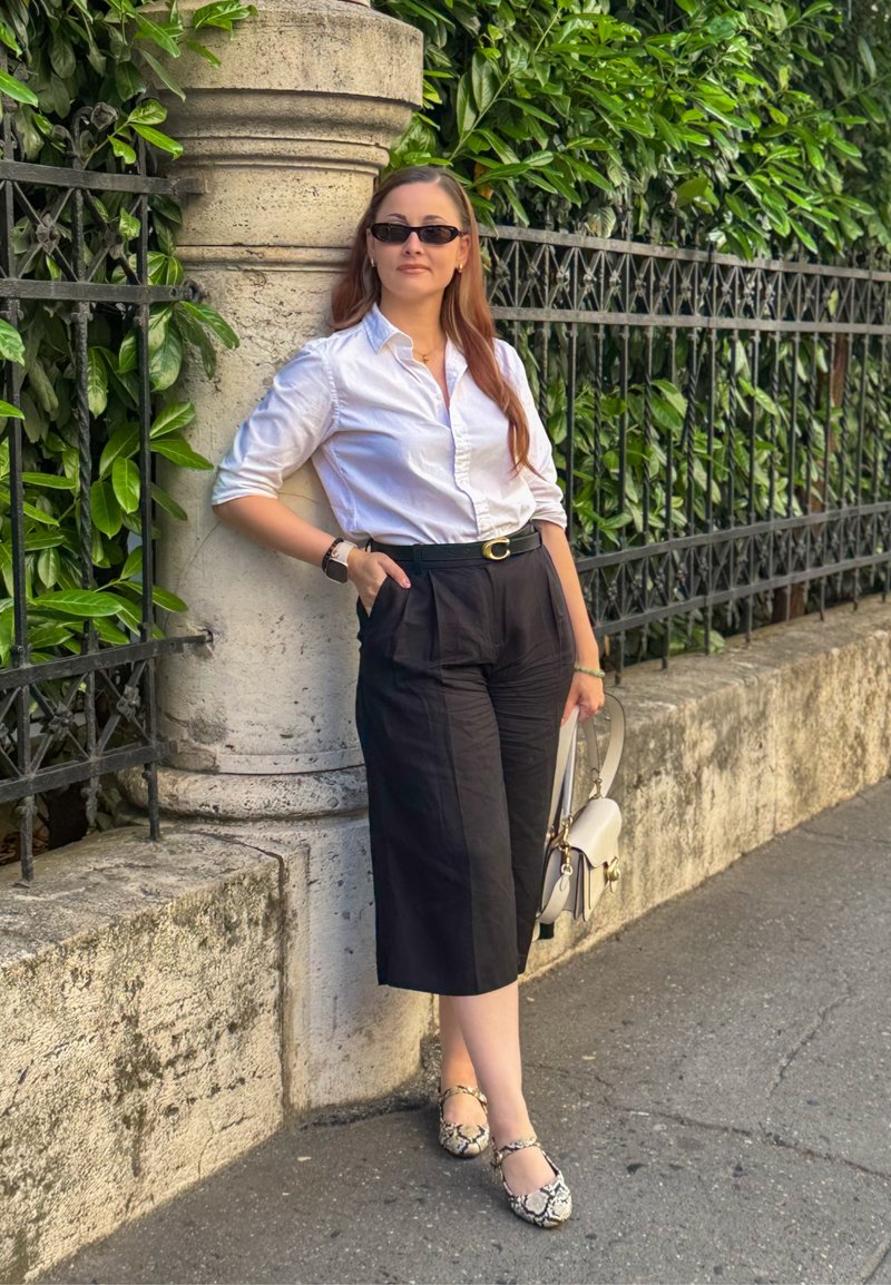 Femme portant des lunettes de soleil, une chemise blanche, un pantalon large noir et des chaussures à motifs, appuyée contre un pilier en pierre avec un feuillage vert et une barrière en fer derrière.