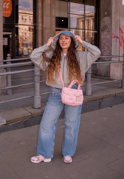 Joven mujer con pelo largo que lleva una sudadera con capucha gris, gorra azul, vaqueros anchos y zapatillas rosas, sosteniendo un bolso rosa de hombro en una calle de la ciudad.