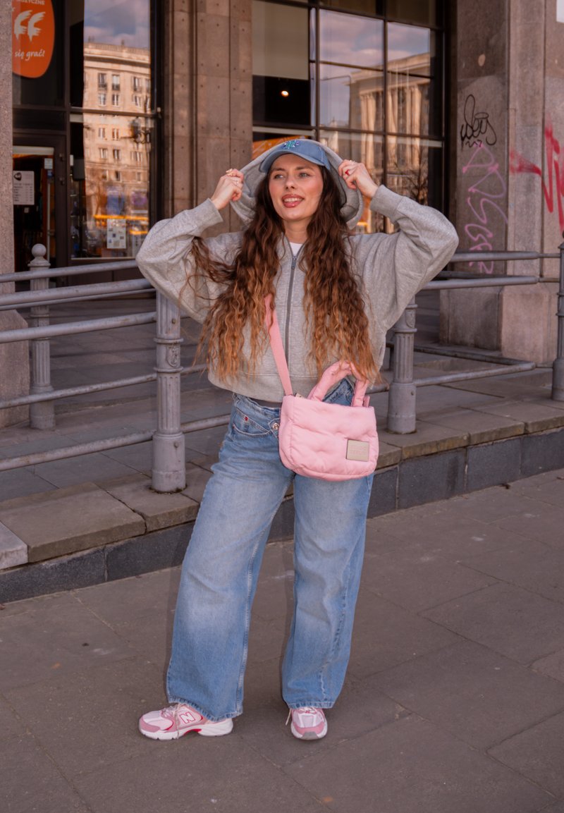 Mladá žena s dlouhými vlasy, která má na sobě šedou mikinu s kapucí, modrou čepici, široké džíny a růžové tenisky, držící růžovou kabelku přes rameno na městské ulici.