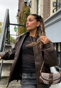 Chaqueta de cuero marrón, camiseta negra y vaqueros oscuros. Sostiene un bolso marrón y beige con estampado y herrajes dorados. Cabello largo y ondulado con reflejos.