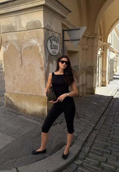 Top negro sin mangas combinado con leggings cortos negros. Lleva un bolso monogramado y usa gafas de sol oscuras. Entorno urbano con adoquines.